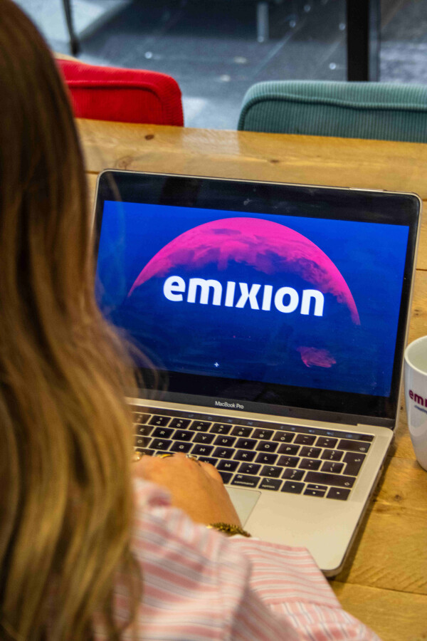 "Vrouw werkt op een MacBook Pro met het Emixion-logo zichtbaar op het scherm, zittend aan een houten tafel met een Emixion-mok ernaast.